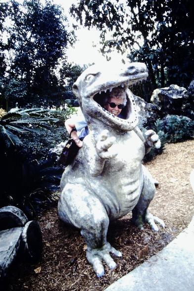 This is a digital scan of a slide from 1982 of an older woman sticking her head through the back of the head of a dinosaur statue.