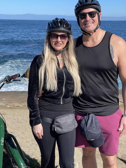 A woman and man wearing bicycle helmets stand alongside one of their rental bikes, their backs to the Monterey Bay.