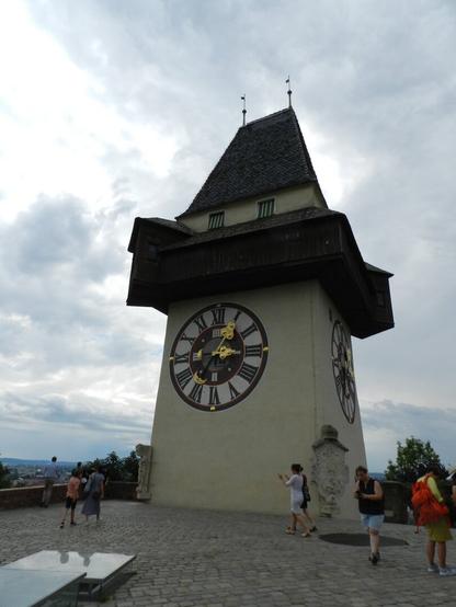 The clock tower in Graz. A small light yellow squared tower with a greyish, slightly pointed, rooftop.