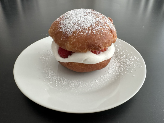 A donut-like bun cut in half and filled with whipped cream, jam, and fresh raspberries, then sprinkled with powdered sugar