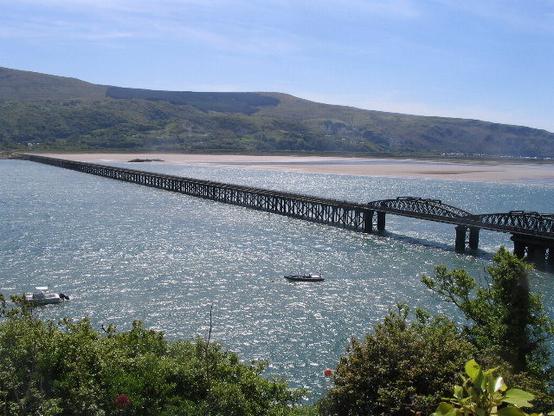 A panoramic view of the Barmouth Bridge, a long wooden viaduct stretching across a sparkling body of water, with hills in the background under a clear blue sky. Boats float gently on the water, and the lush greenery of the coast is visible on both sides.