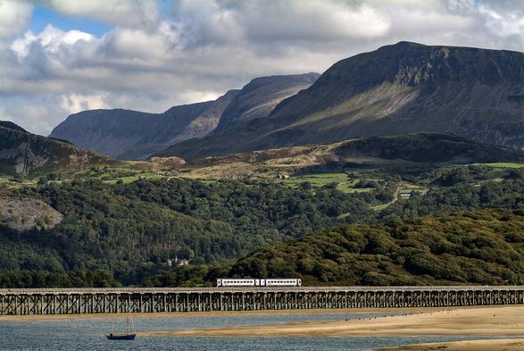 A picturesque scene of the Barmouth Bridge with a train crossing over. The bridge is framed by the rugged Welsh mountains in the background and a serene beach in the foreground. The bridge's wooden structure stands out against the natural beauty of the surrounding landscape.