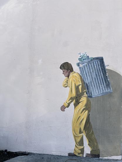 A mural depicting a city worker wearing yellow coveralls and hauling a metal garbage can on his back with an alien poking its head out.