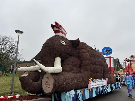 A parade float designed to resemble a large brown mammoth with white tusks and a hat, with banners and decorations, surrounded by people in festive attire.