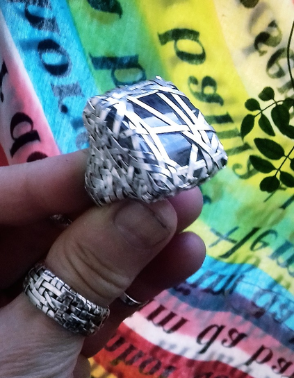 hand holding a ring handcrafted in silver strips woven into an intricate pattern encasing a moss agate stone, in the background a fabric from Bahia, Brazil with the rainbow colours