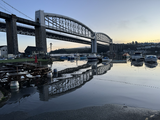 Brunel’s rail bridge spanning the flooded Tamar river as sun starts to rise hiding just behind the hillside on the opposite bank. Foreground with pub bar tables sitting in the high water
