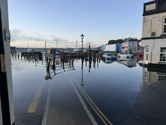 Tamar river so high is come up the slipway on the left and covered the road in front of me all the way to the restaurant on the opposite side. Footpath and yellow lines painted on the road submerged below the water. Several cars nearby also with their wheels submerged by several inches.