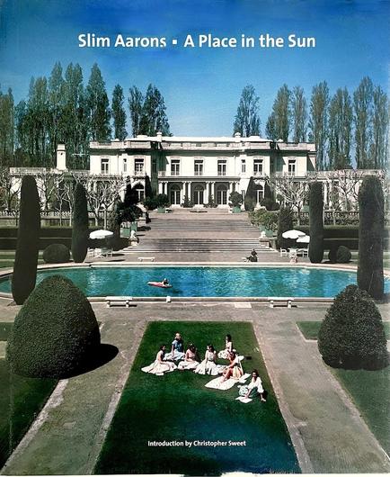 A photography book entitled Slim Aarons: A Place in the Sun. Front cover shows a large mansion in the background. We are looking down on the formal gardens and a long oval swimming pool on a summer day. A group of 7 ladies in summer dresses a sitting on the grass in the foreground.
