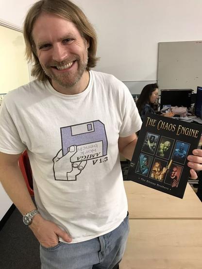 A photograph of me, a blond-ish long-haired middle-aged white guy with a beard in a white t-shirt and jeans, clutching a copy of The Chaos Engine for the Amiga. In a bizarre coincidence, I'm wearing my Workbench 1.3 t-shirt with the Amiga boot screen on it.