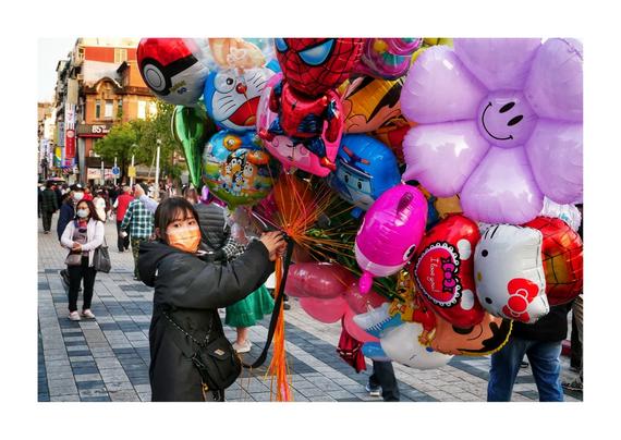 The image shows a person standing on a busy street selling a colorful array of character balloons. The balloons feature various designs, including Spider-Man, Hello Kitty, and Doraemon, among others. The seller is wearing a black jacket and an orange face mask, likely as a health precaution. The background suggests this might be in an Asian city, with a crowd of people walking by, some also wearing face masks. The setting appears lively and is possibly a shopping or commercial area, indicated by storefronts and signage in the background.