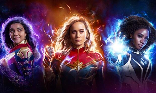 Iman Vellani as Ms. Marvel, Brie Larson as Captain Marvel and Teyonah Parris as Monica Rambeau. All in costumes looking FIERCE with blue, purple, yellow electricity and flames behind them.
