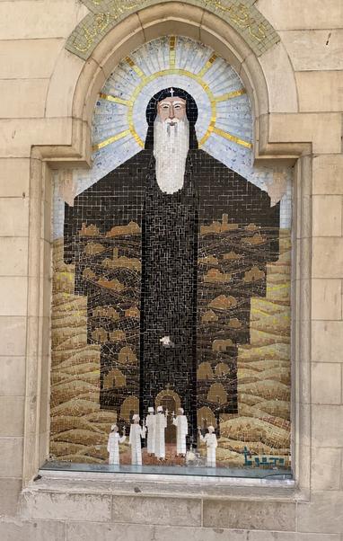Large mosaic built into an outside wall. It shows a white-bearded old man in hooded brown robes. There’s a white cross on his hood and what looks like a halo behind him. location: Cairo, Egypt, Old Coptic Christian Quarter