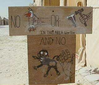 In front of tents in a sandy environment, a series of plywood signs use rebus-style cartoon art to forbid chicken-choking, meat-beating, and monkey-spanking.