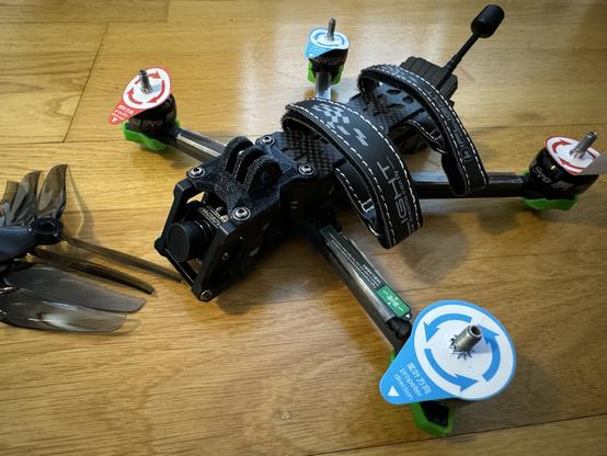 Photo, a 5” freestyle FPV drone on a wooden floor. The motors still have little labels describing what direction they turn in, the props sit next to it ready to be put on.