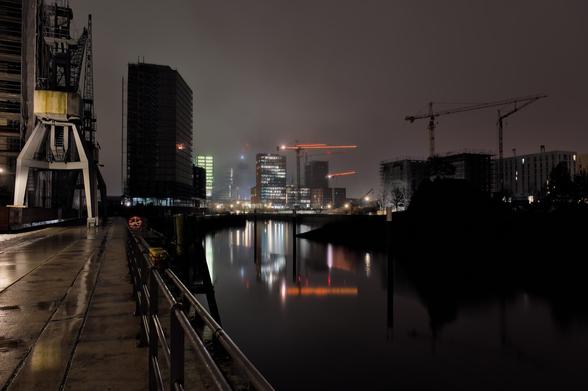 In the center and on the right side of the picture you can see the cranes of a construction site. The cranes in the middle have fairy lights in their booms. On the left side you can see buildings and an old harbor crane. Some of the windows are lit. A former harbor basin can be seen in the lower half of the middle, in which the scenery is reflected. The picture was taken at night.