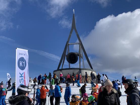 The clock at the top of the Kronplatz, ringing.
There’s a crowd waiting. The sky is slightly cloudy in the picture, however the general weather was gorgeous. There is a banner from Josk on the left side, a banner for K1 beginner level kids for skiing.