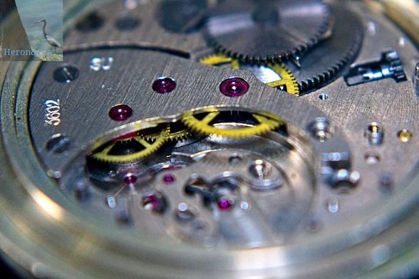 A macro view photograph of the internal clockwork of a pocket watch, centered on three ruby bearings showing their tops on steel casing and brass gears underneath.