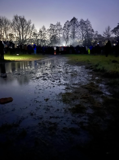 Dusk is setting in the sky, foreground is dominated by a still body of water, filled with plants, reflecting the darkening sky. To the left in the near distance, a street light highlights the green field in which many silhouettes of people can be seen, dotted with some in reflective clothing