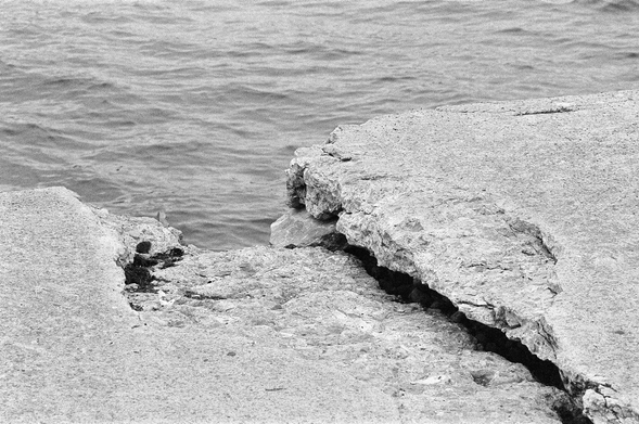 A black and white photograph showing a large open crack in a concrete quay, a large piece of concrete is missing towards the water’s edge.