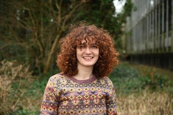 Portrait photo of Anastasia, with sholder-long dark red curly hair, smiling in the camera.