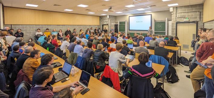 Picture of the said devroom, with all seats taken and many people standing to listen to the ongoing talk