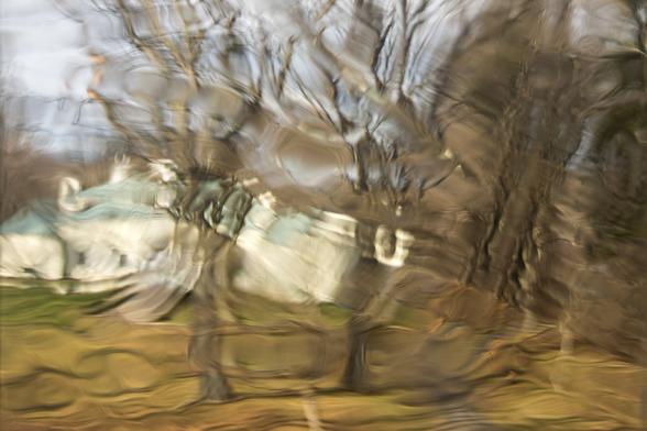 A color photograph of a museum building and a few large, bare trees on a green lawn. Taken through an old glass window, the view is severely rippled and distorted, almost looking melted.