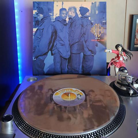 A clear tan vinyl record sits on a turntable. Behind the turntable, a vinyl album outer sleeve is displayed. The front cover shows 4 members of Jodeci posing in coats.  3 are looking away, while 1 member looks at the camera.