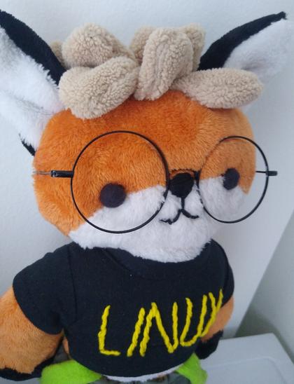 picture of... of.... xenia plushie???? wow!!! yeah!
xenia plush, the standard orange fox with white lower mouth fur, white inner ears, green visible suspenders and a black shirt that says LINUX. she is wearing wide circular glasses as well
:3