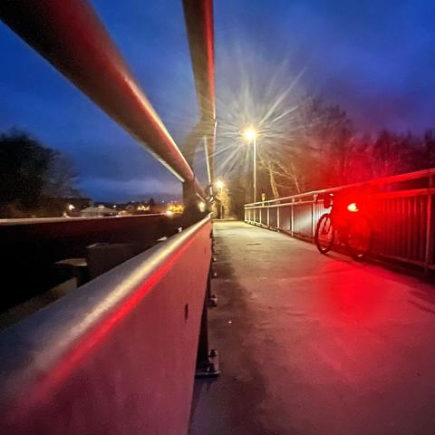 Eine Brücke mit gemeinsamem Fuss- und Radweg in der Dämmerung mit Straßenlaternen und einem abgestellten Fahrrad an der Seite.