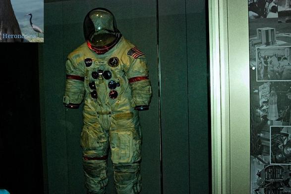 An indoor, dimly lit photograph of a space suit in a glass museum display. The helmet’s front is clear, with a black cap reaching from the back. The ring connecting the neck to the collar is bright red. The suit is white. The US flag is on the left sleeve, and both sleeves have a pocket and a red stripe, ending in red and blue rings where the gloves would attach. The center of the suit has the blue NASA emblem on the left, the name SHEPARD on a patch in the center, a circular emblem for the Apollo 14 mission. There are six connectors mounted on the front of the suit, which are circular and metallic blue or red. Straps help hold the suit tight. The legs have pouches and red stripes. The was behind is neutral gray.