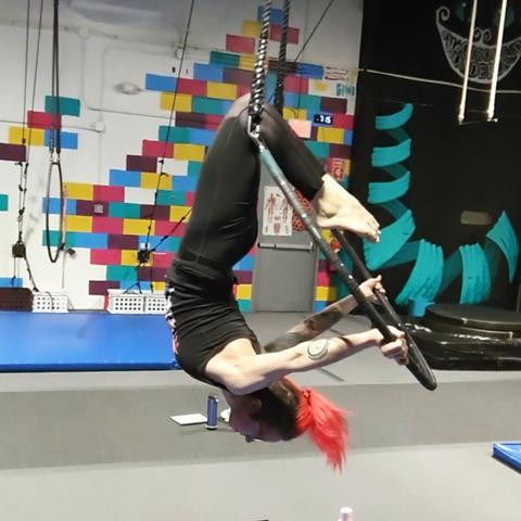 A white woman with red hair hanging from her knees on the top of a lyra or aerial hoop. She is wearing all black and her arms are pushing the hoop behind her.