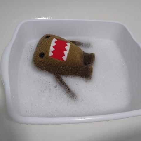 Domo-kun, a small square brown furred creature, with a big open red mouth and two sets of identical sharpened teeth (4 on the top, 4 on the bottom), laying peacefully on top of a soapy bubble bath in a small wash square bucket.