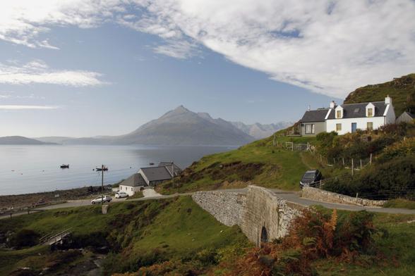 Eine kleine Straße führt auf eine Bucht. Ein paar Häuser stehen am Hang. Im Vordergrund eine Brücke aus Stein, Farne und Gräser wachsen auf Wiesen. Auf dem Wasser liegen Boote vor Anker, im Hintergrund Berge. Der Himmel ist blau, Schleierwolken ziehen am rechten Rand auf. 

A small road leads to a bay. A few houses stand on the slope. In the foreground, a stone bridge, ferns and grasses grow on meadows. Boats are anchored on the water, mountains in the background. The sky is blue, veil clouds are gathering at the right edge.