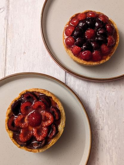 Two individual tarts with raspberries and cherries on top, sitting on two seperate plates on a white wood background.