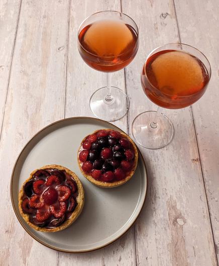 Two individual tarts with cherries and raspberries arranged decoratively on top sat side by side on a plate, and two Nick and Nora glasses with pinkish-red clear drinks in them, all sat on a white wood background.