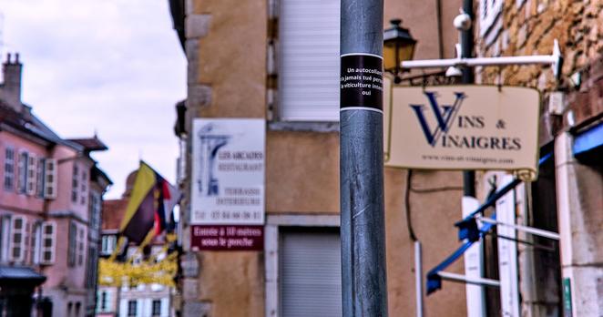 Un autocollant noir avec écriture blanche. Il est collé sur un montant métallique d'un panneau de signalisation de circulation en centre du village d'Arbois. Juste derrière on aperçois une enseigne « Vins et Vinaigres » d'une boutique et le décor de fond est composé des bâtiments en pierres anciennes aux couleurs jaune terre et rose passé sur crépis avec des drapeaux pendants des petits mats de premiers étage.