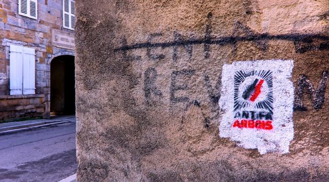 Sur un mur en crépit ancien de le cœur de village à un angle de rue, une peinture rectangulaire format portrait au pochoir. Sur un fond blanc deux bouteilles de vins rouges et noires surplombent le slogan Antifa (en couleur noir) Arbois (couleur rouge)