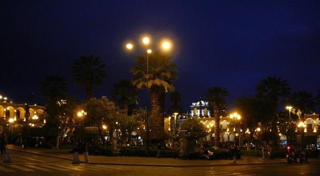 Foto general de la Plaça d'Armes. Al centre hi ha uns jardins amb palmeres i gent i al fons hi ha dos laterals porxats.