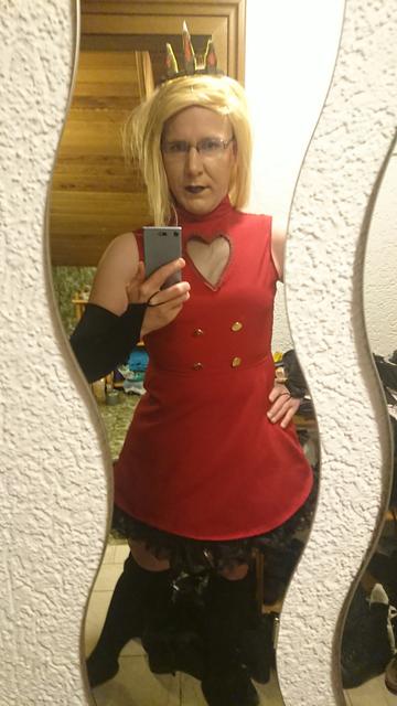 A photo of myself wearing a red, sleeveless dress that reaches till half the upper legs. The "skirt" part of the dress has a black petticoat below it. The dress has four messing buttons around the stomach area and a heart shaped cutout with transparent material above the beast area. I'm wearing a long, blonde wig with bangs styled out of the way.
Additionally I'm wearing knee length socks and fingerless gloves that reach till above the elbows.