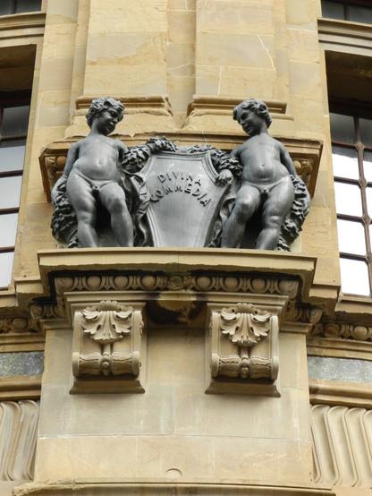 A stone carving on a building featuring two figures holding a shield inscribed with the text “DIVINA COMŒDIA.”