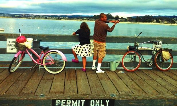 A girl in a white and black skirt and black top stands on Monterey's Wharf II alongside her dad, who is dressed in tan shorts and a brown top. Each has their bicycle alongside them. Hers is a pink bicycle, with a dark pink helmet and a white jacket in a front basket. His is a black bicycle with tan cargo pouches on the rear. The dad is about to cast out his fishing line. The girl is leaning on the wharf railing, looking over the Monterey Bay.