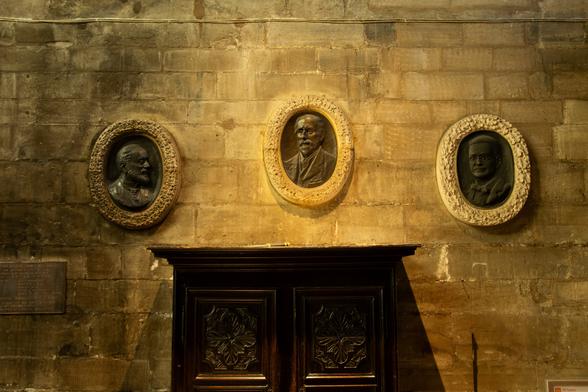 Wall of an interior of a church, somewhat dimly lit, showing three ovals with bas-reliefs of men's heads. Below it part of a wooden cabinet.