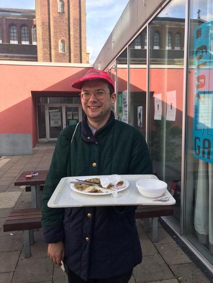 Ein junger Mann steht auf einer Terasse vor einem Gebäude. Er trägt Dreitagebart, eine Brille, eine grüne Jacke, eine verblichene rote Kappe mit der Aufschrift "Mamba" und lächelt in die Kamera. In der Hand hält er ein typisches Mensatablett, auf dem eine kleine Schüssel und ein leergegessener Teller stehen.