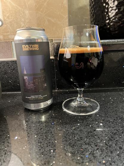 A can of Evil Twin Brewing Christmas Eve in a New York City Hotel Room - Royal Suite Edition beer. The label features the New York City skyline in shades of dark purple and black. A tulip glass is to the right half full of the dark-as-night brew.