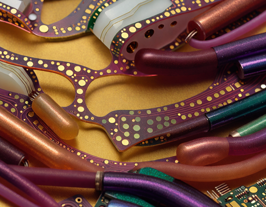 Close-up of flexible circuit boards, with pads arranged like polkadots, and colorful insulated wires on a golden background, reflecting light on shiny surfaces.