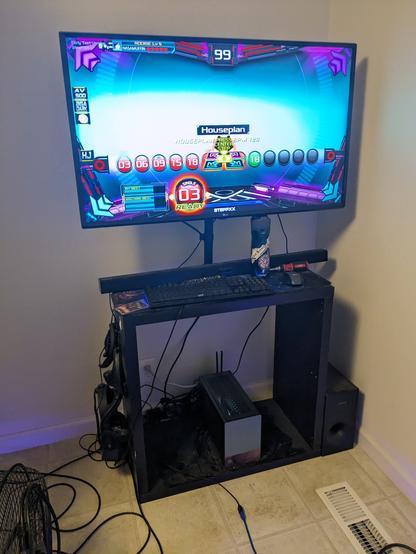 A 2x2 Kallax cube shelf with the shelves removed - so it's just a big square frame - has a decently sized TV mounted to it, a sound bar and keyboard laid on top, and a comically small jade green desktop PC sitting inside. The TV is showing the chart difficulty selection screen in StepPXX, an illegal Pump It Up clone.