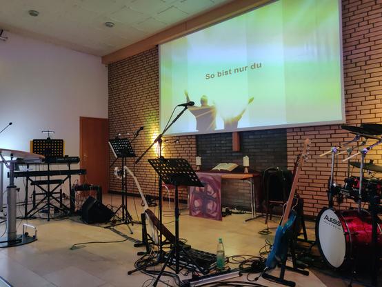 "Bühnen"-Bereich eines modernen Kirchengebäudes mit Band-Setup: links Keyboards, in der Mitte Mikrofone, Notenpulte und Gitarre/Bass, rechts Schlagzeug. Abendmahlstisch, darüber eine Leinwand mit Figuren mit erhobenen Armen in Gegenlicht, großer Text "So bist nur du".