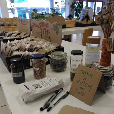 A selection of seed packets in trays on a desk, with glass jars of seed next to a sign saying "Take a pinch or two"