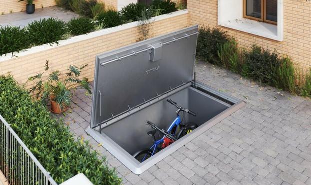 A paved front garden with a large metal storage area sunk into the ground, the lid open, and two bicycles inside.