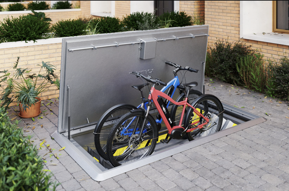 A metal storage container fitted into the ground with the lid open and two bikes lifted up to ground level from inside.